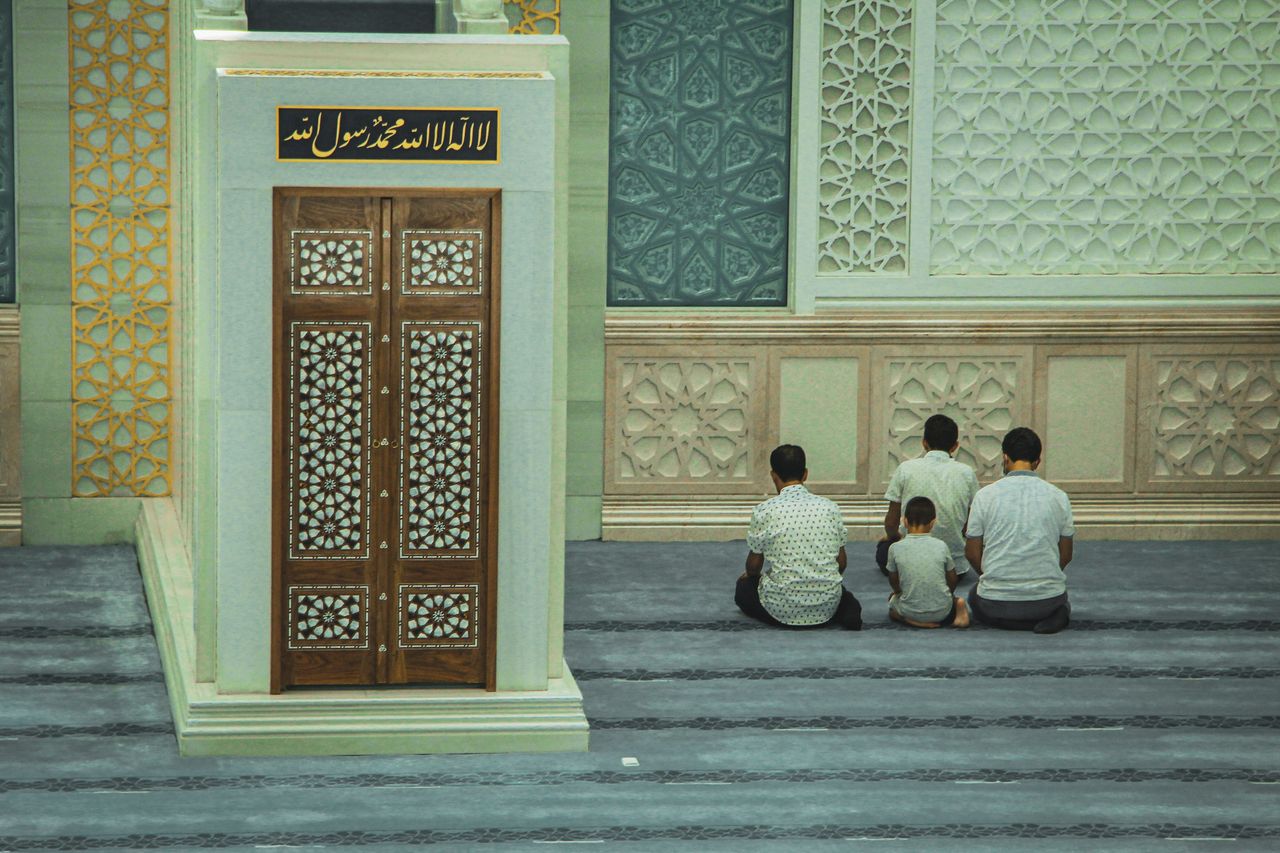 Group of men and a child pray inside a mosque with intricate wall decorations.